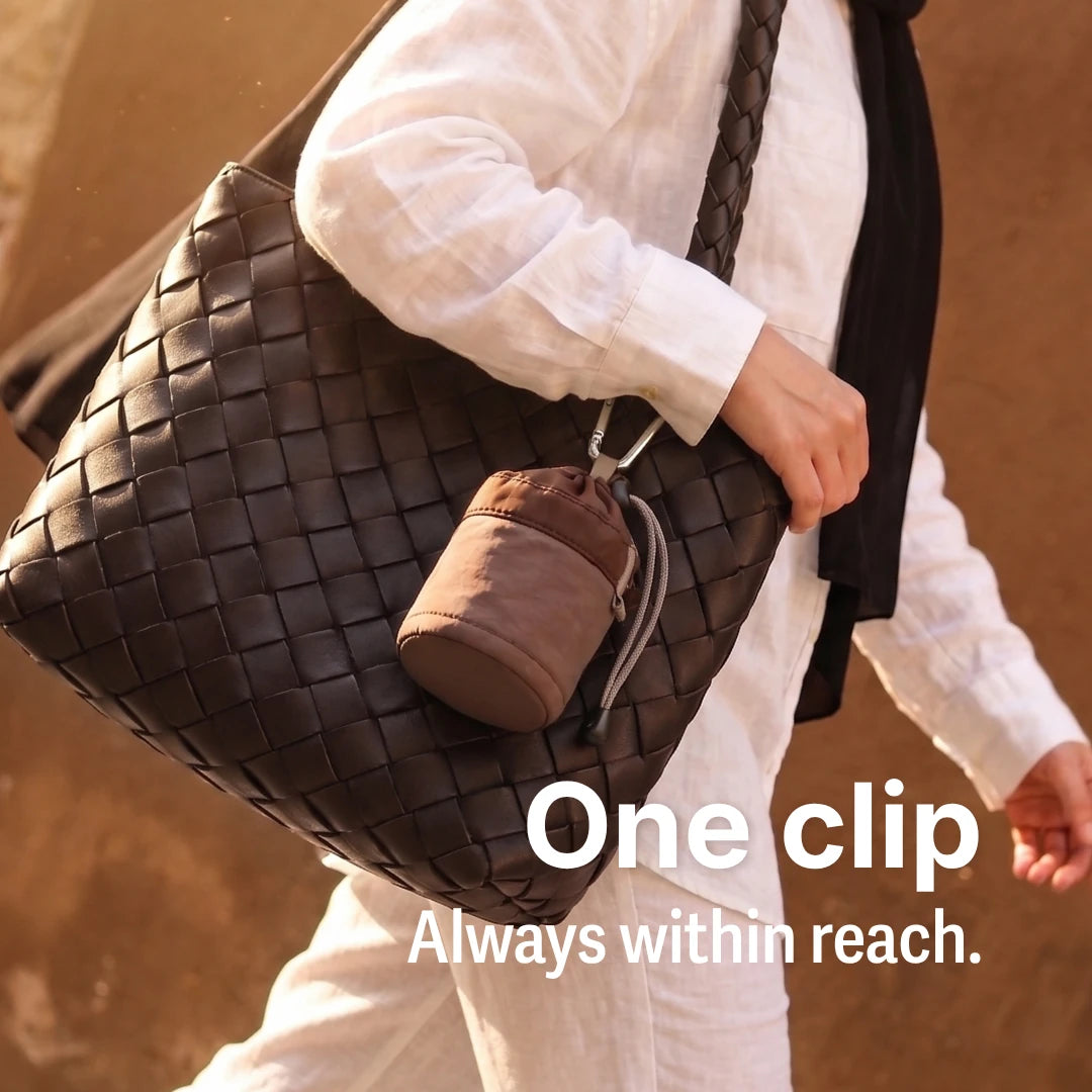 Lifestyle shot of a person in a white outfit carrying a large woven dark brown leather shoulder bag with SalahWear cuppa cocoa brown pouch clipped to it. The text shown is 'One clip, Always within reach.